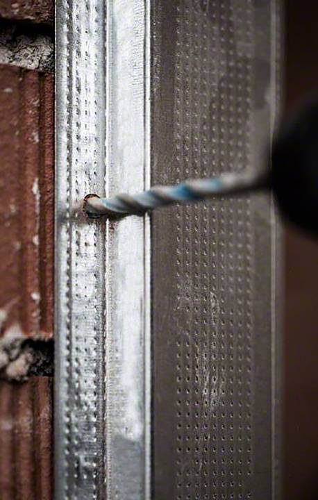 Wiertło przenika przez profil metalowy na ścianie z cegły.