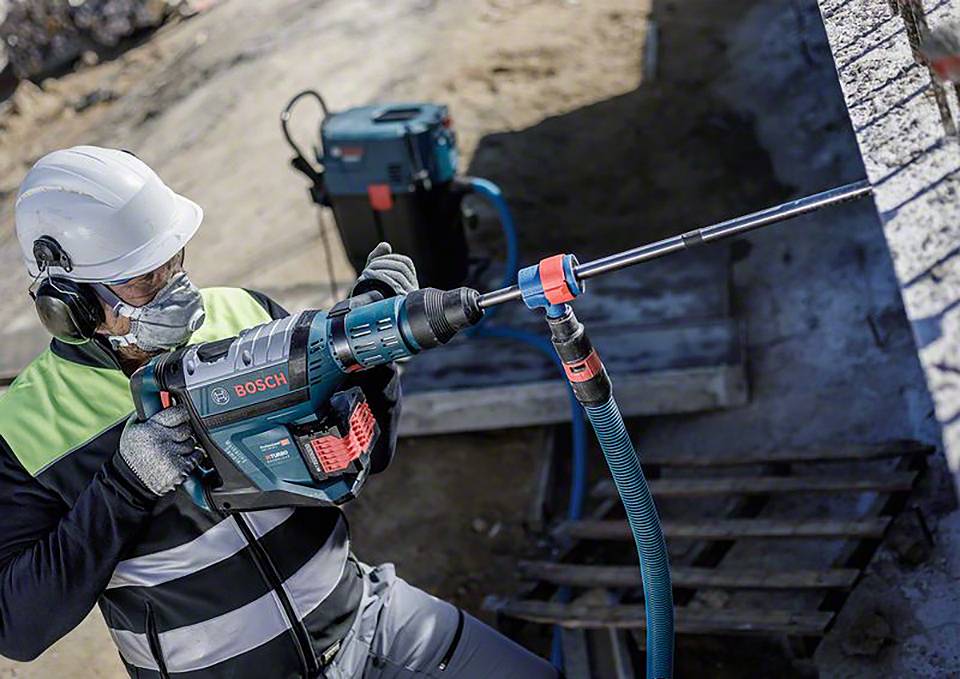 Osoba w odzieży ochronnej i kasku wierci otwór w betonowej ścianie za pomocą młotowiertarki Bosch na placu budowy.