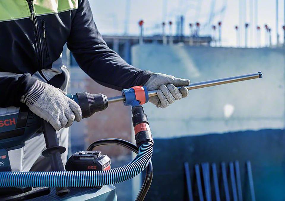 Osoba ubrana jest w rękawice robocze i używa elektrycznej wiertarki. W tle widoczne są struktury budowlane.