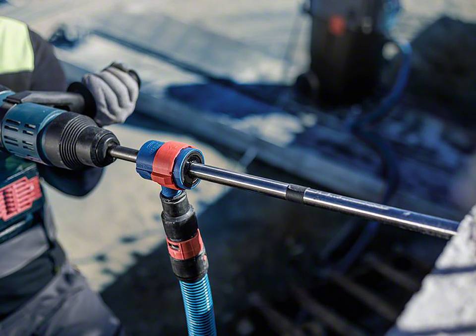 Osoba używa elektrycznej wiertarki do wiercenia w betonie. Scena przedstawia roboty budowlane w świetle dziennym.