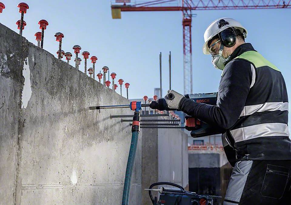 Robotnik budowlany w kasku ochronnym i nausznikach wierci w betonową ścianę na terenie budowy. W tle widać dźwig.