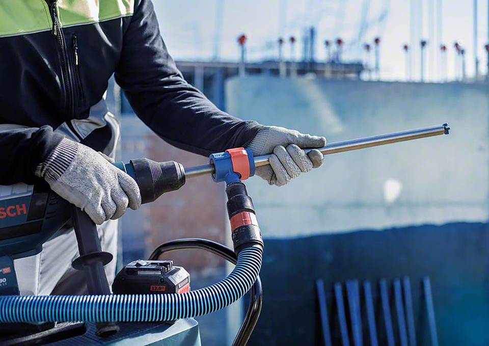 Robotnik używa wiertarki na placu budowy. Ma na sobie rękawice robocze i odzież ochronną. W tle widać budynki.