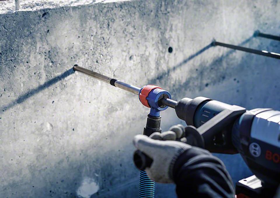 Rzemieślnik wierca otwór w betonowej ścianie za pomocą elektrycznej wiertarki. W tle widać jasne światło.