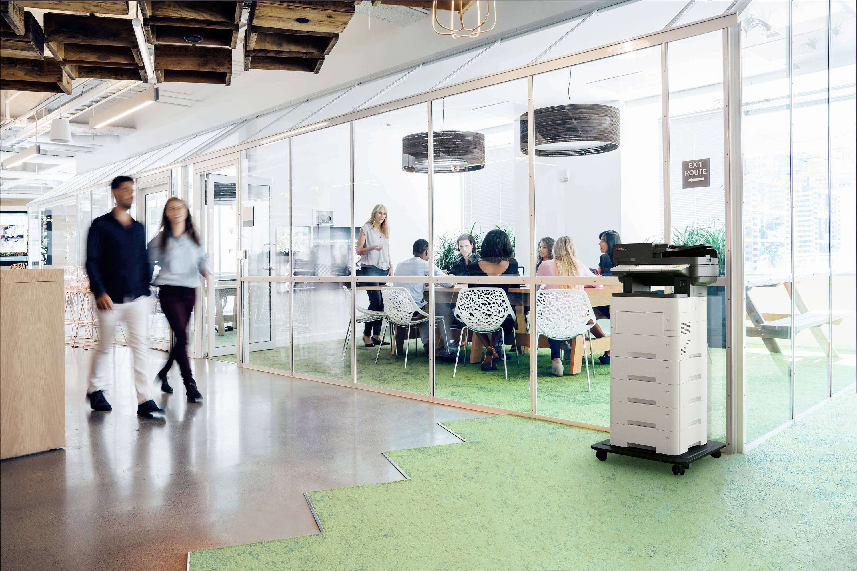 Środowisko biurowe z przeszklonymi ścianami, wewnątrz ludzie siedzą na spotkaniu. Dwie osoby przechodzą obok wielofunkcyjnego drukarki.