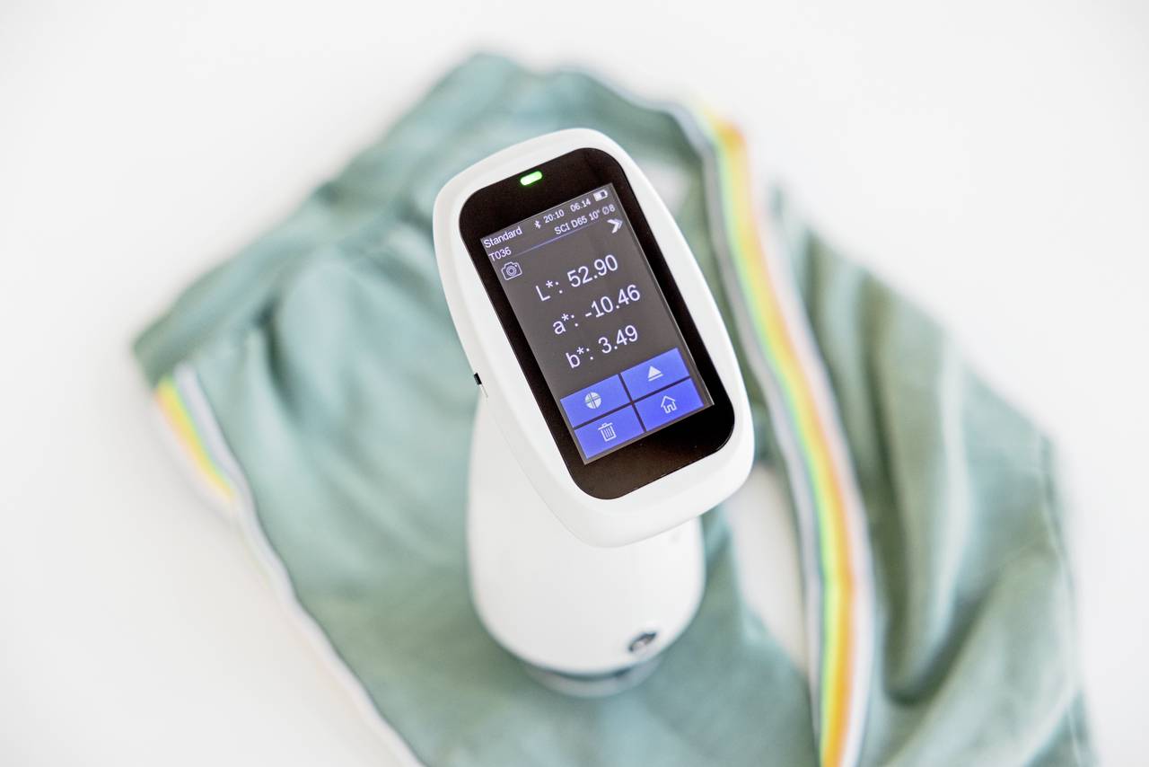 Urządzenie ręczne z wyświetlaczem pokazuje wartości kolorów. Leży na składanym, turkusowym kawałku tkaniny w kolorowe paski.