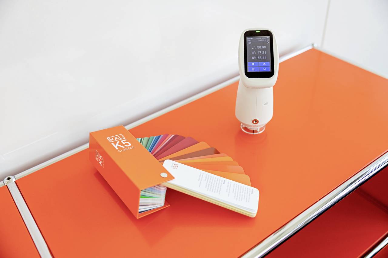 Miernik koloru umieszczony obok wachlarza z próbkami kolorów na pomarańczowym stole. Urządzenie wyświetla wartości pomiarowe na wyświetlaczu.