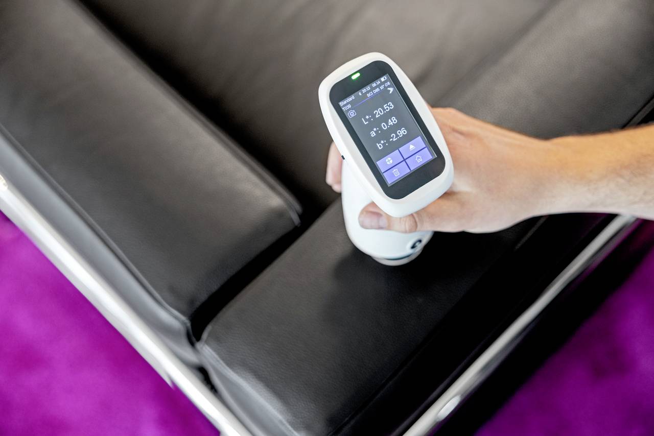 Ręka trzyma cyfrowy miernik koloru nad czarną skórzaną sofą. Urządzenie wyświetla pomiary na wyświetlaczu.