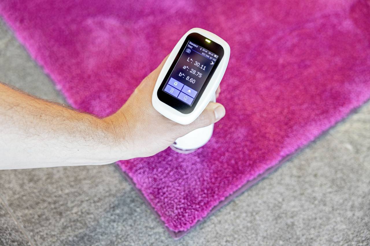 Osoba trzyma cyfrowy miernik koloru nad różowym dywanem na szarej podłodze. Wyświetlacz urządzenia pokazuje dane pomiarowe.