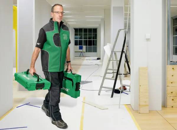 Pracownik w zielono-czarnym mundurze niesie dwie zielone skrzynki z narzędziami przez jasno oświetlone, nieukończone pomieszczenie biurowe, w którym stoją drabiny i leżą materiały.