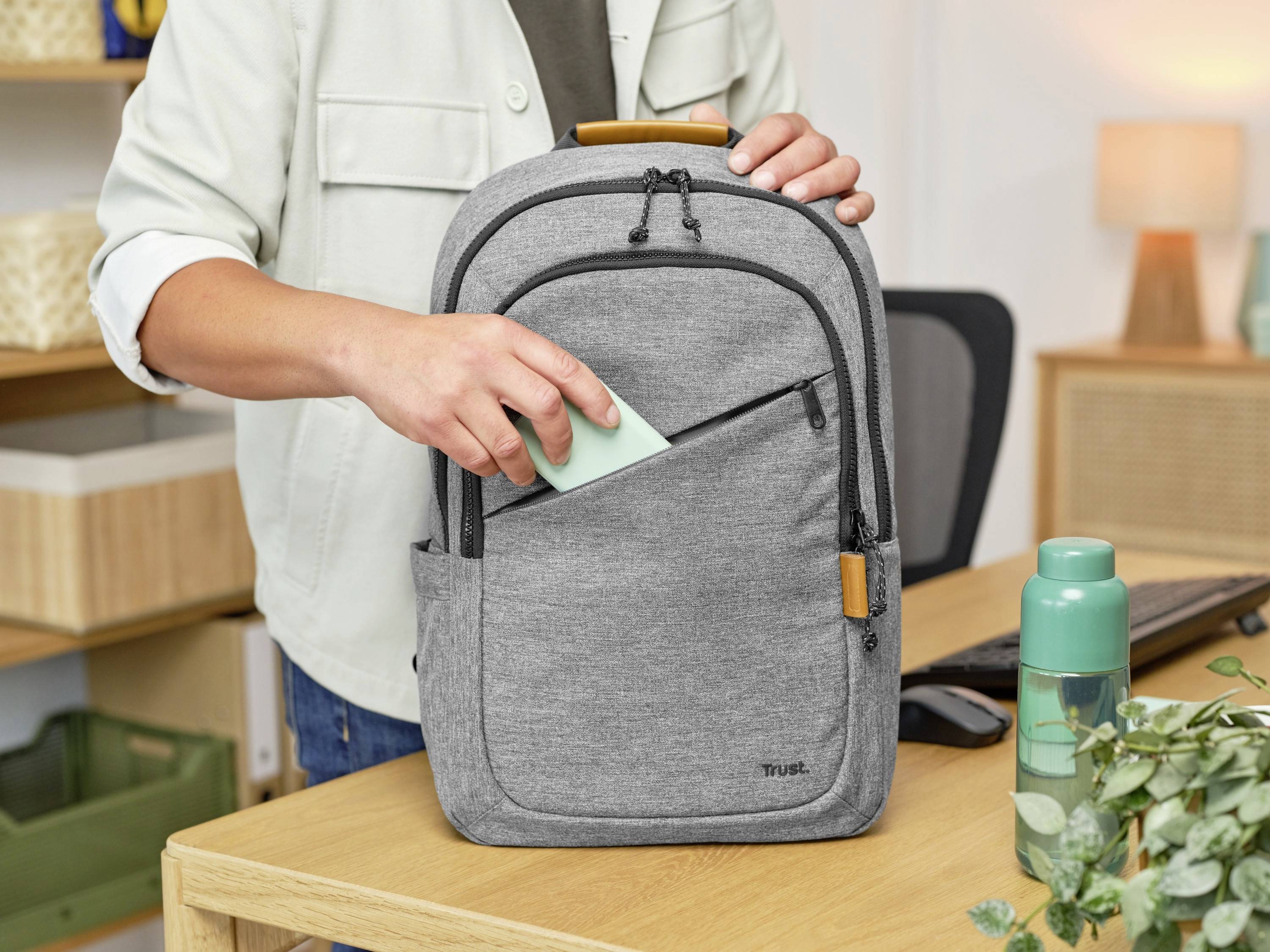 Osoba wkłada zieloną kartkę do przedniej kieszeni szarego plecaka, który leży na biurku obok butelki wody.