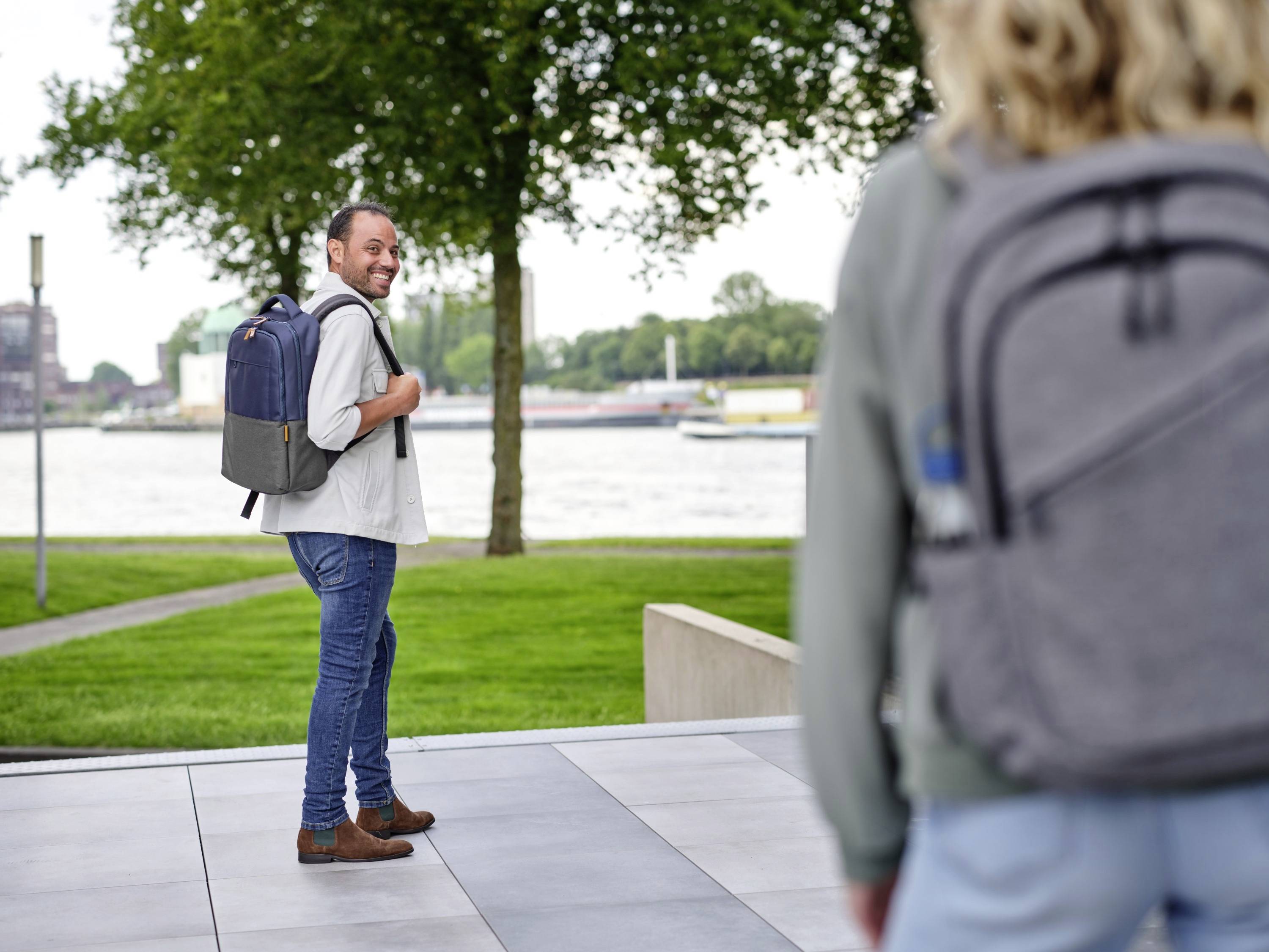Mężczyzna z plecakiem, uśmiechając się, ogląda się za siebie, spacerując w parku z widokiem na rzekę. Na pierwszym planie niewyraźna postać.