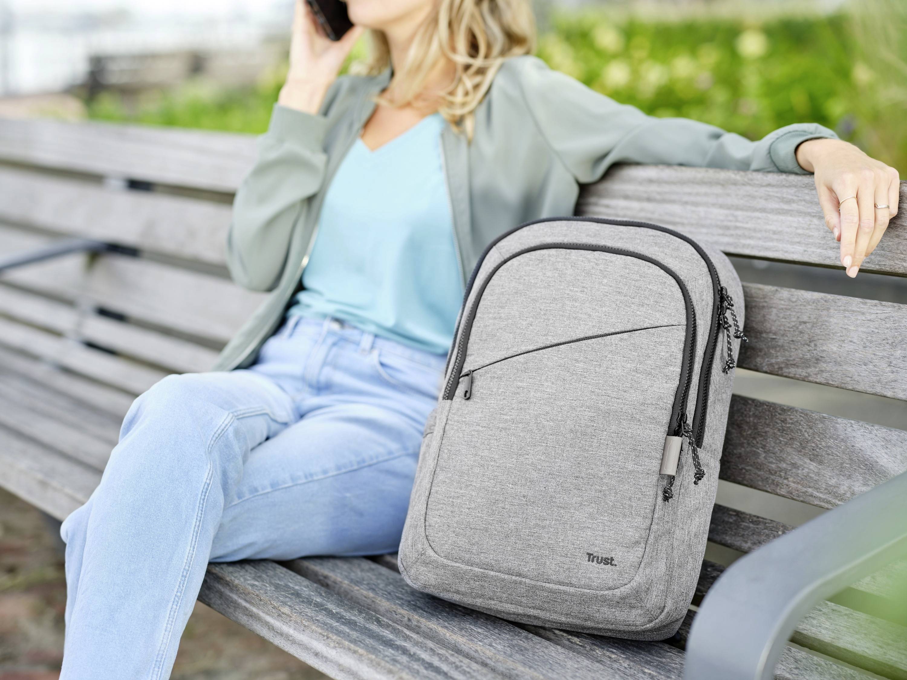 Kobieta siedzi uśmiechnięta na ławce w parku i rozmawia przez telefon, obok niej stoi szary plecak.