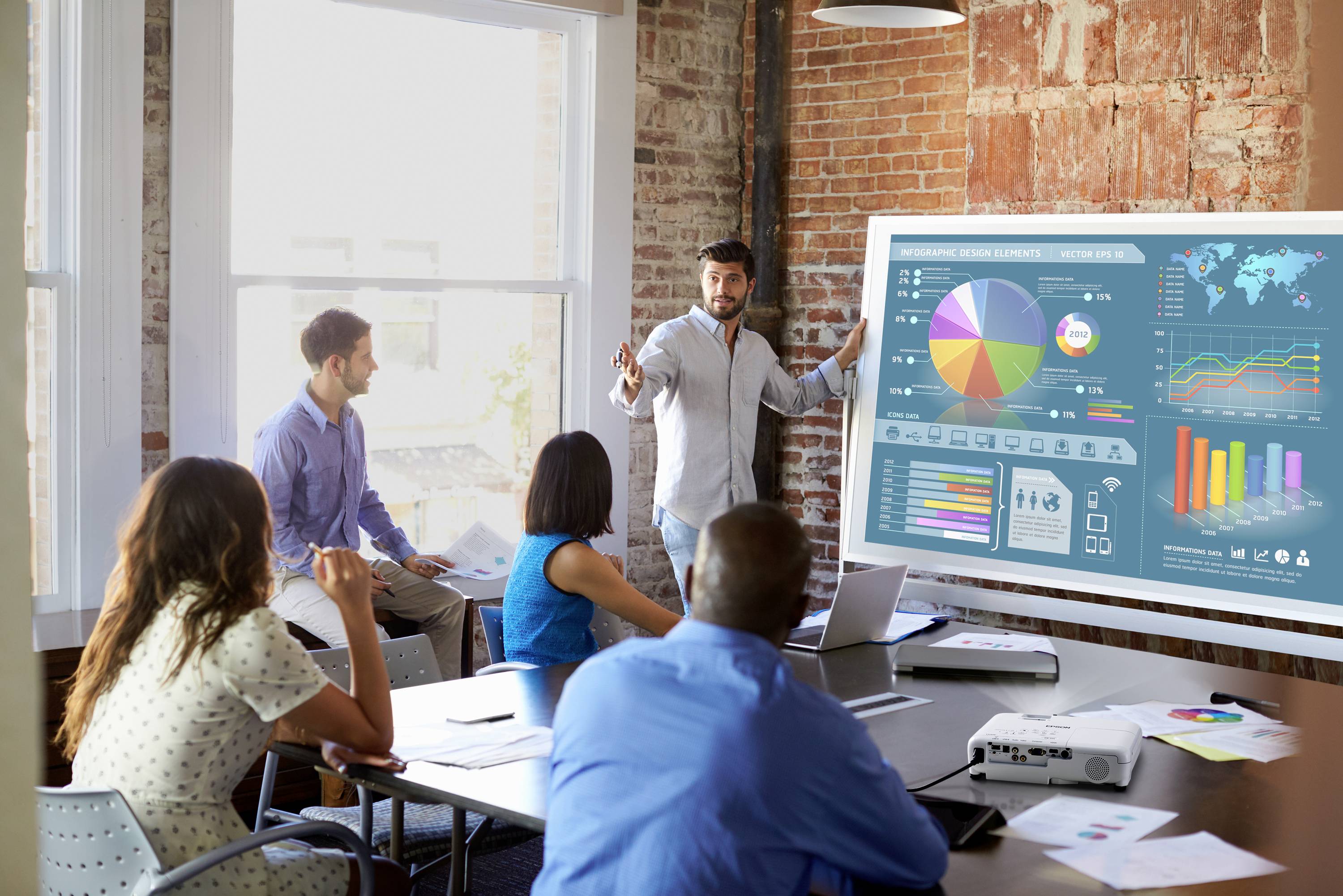 Mężczyzna prezentuje w sali konferencyjnej przed kolegami, wskazuje na diagram z kolorowymi wykresami i statystykami wyświetlanymi na dużym ekranie.