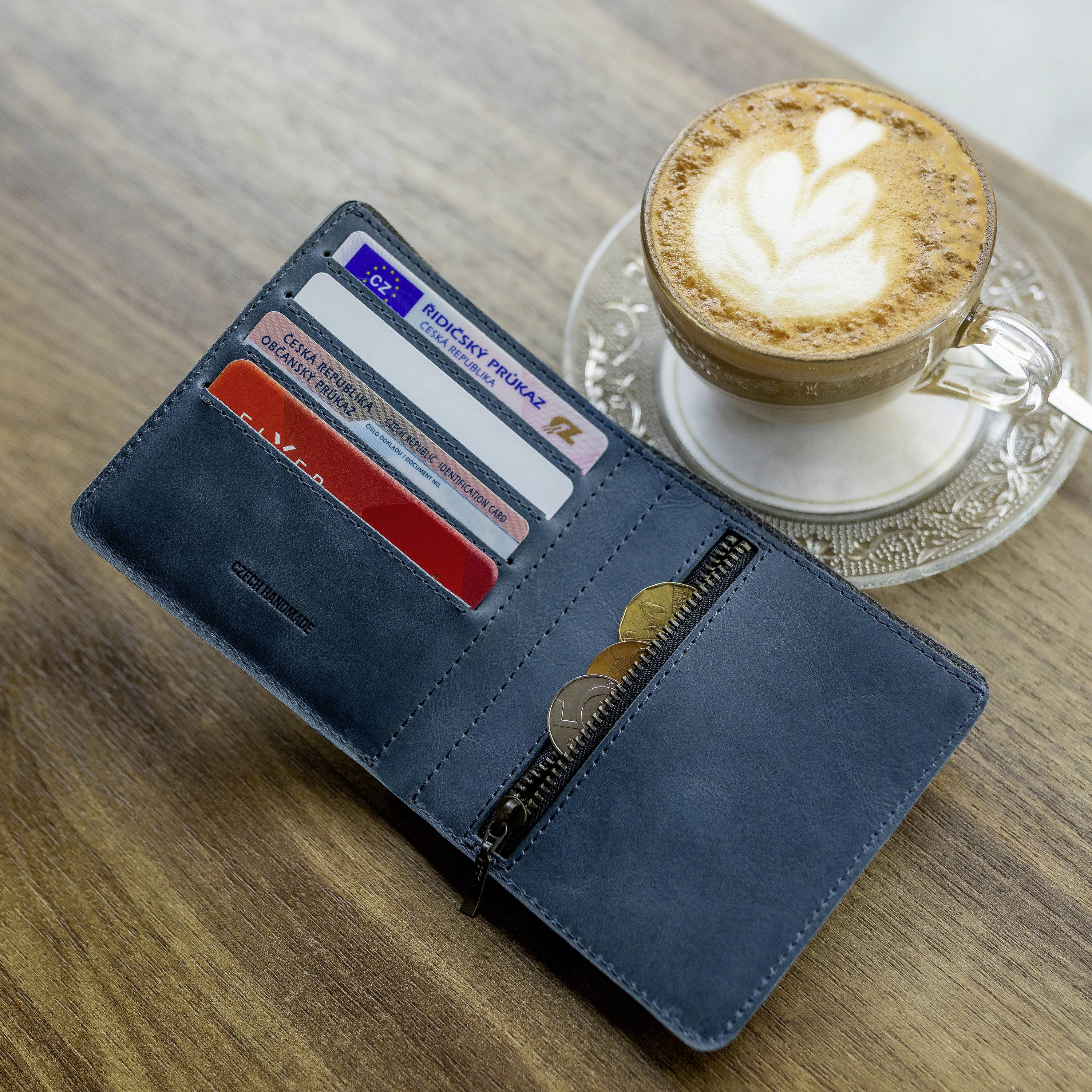 Otwarta niebieska portmonetka z kartami kredytowymi i monetami leży na drewnianym podłożu. Obok stoi filiżanka cappuccino.