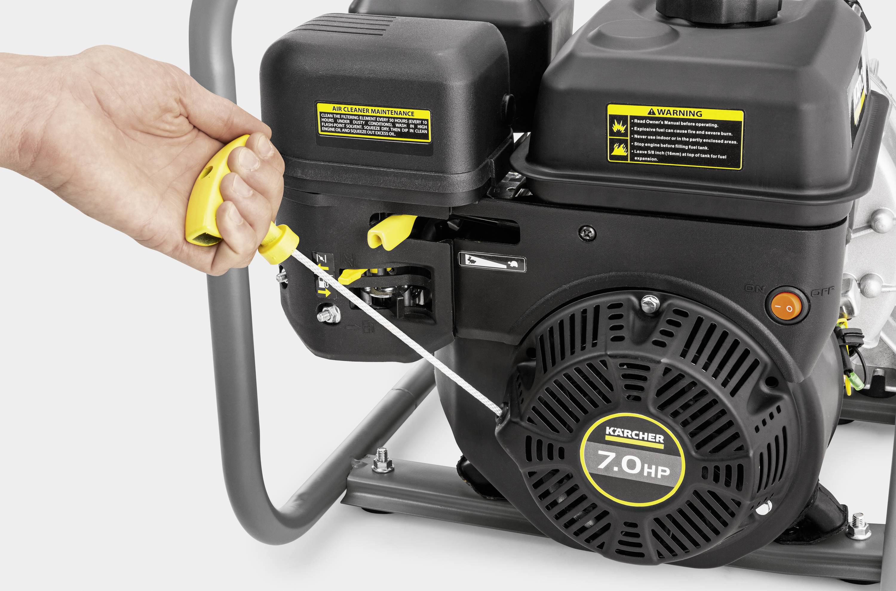 Jedna ręka pociąga linkę rozruchową silnika o mocy 7,0 KM maszyny czyszczącej. Na obudowach silnika umieszczone są ostrzeżenia.