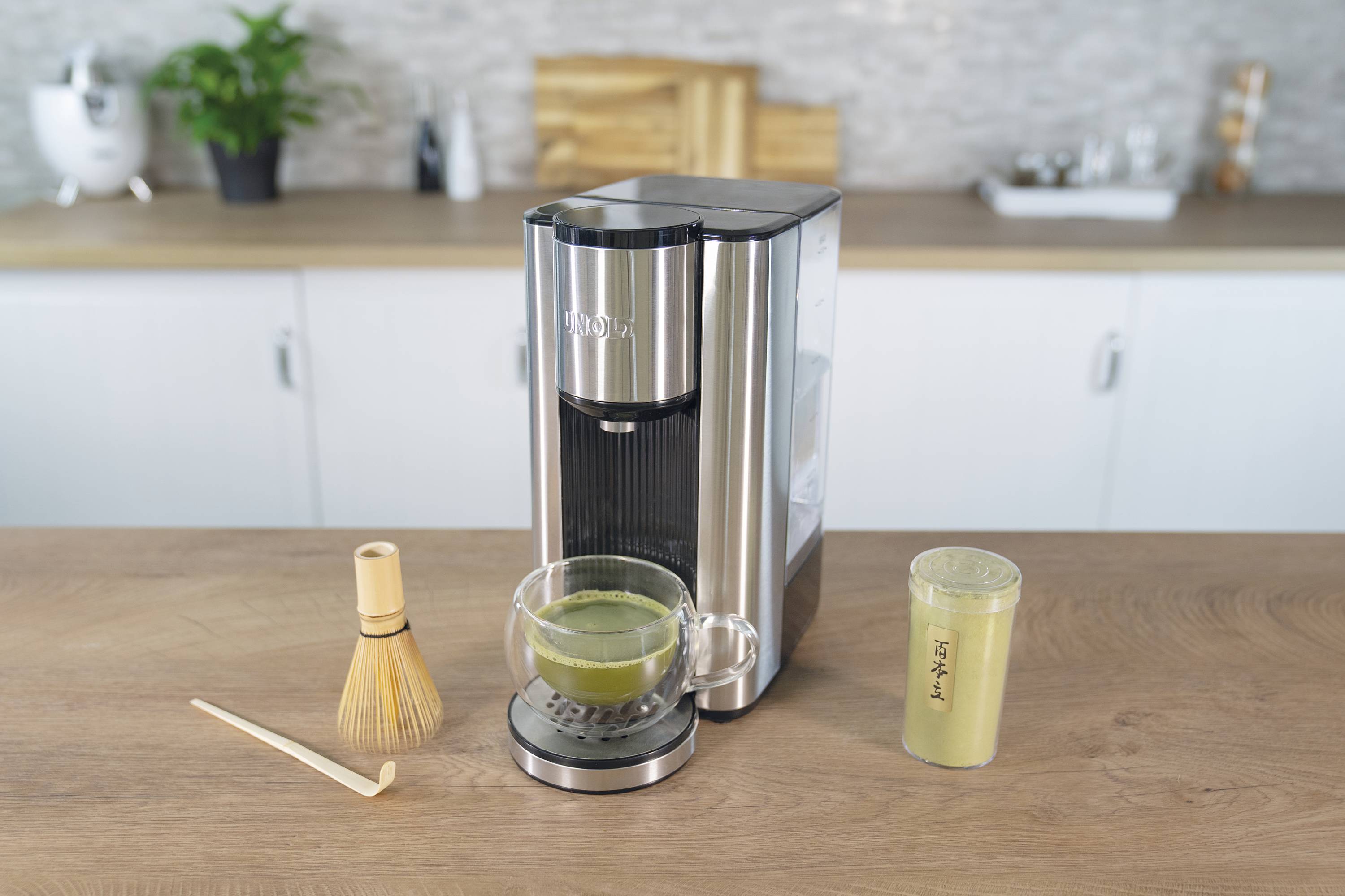 Maszyna do matchy stojąca na blacie przygotowuje świeżą herbatę matcha. Obok leży bambusowa trzepaczka i szklanka z proszkiem matcha.