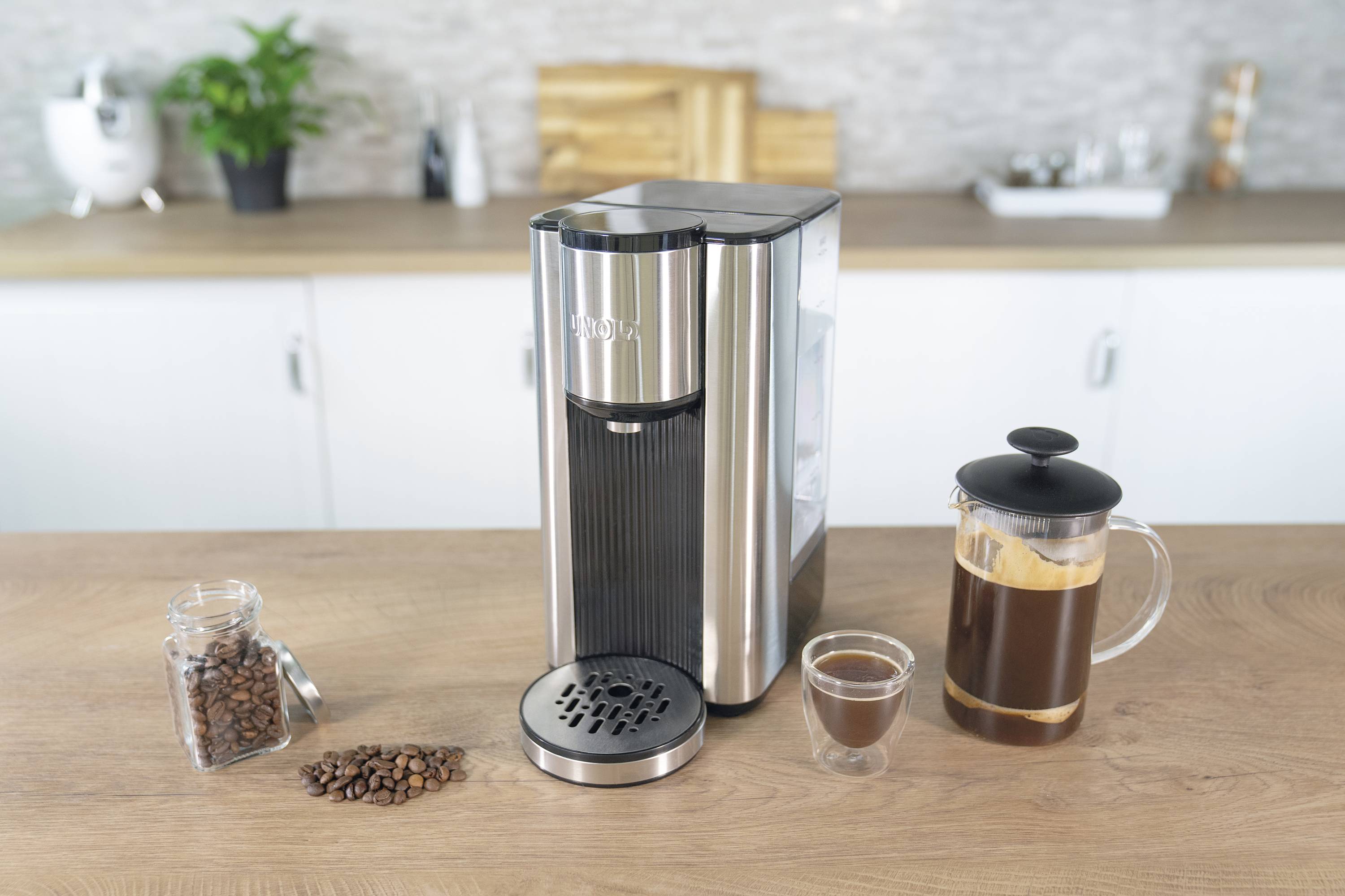 Srebrny ekspres do kawy stoi na blacie kuchennym, otoczony szklanką kawy, ziarnami kawy i zaparzaczem.