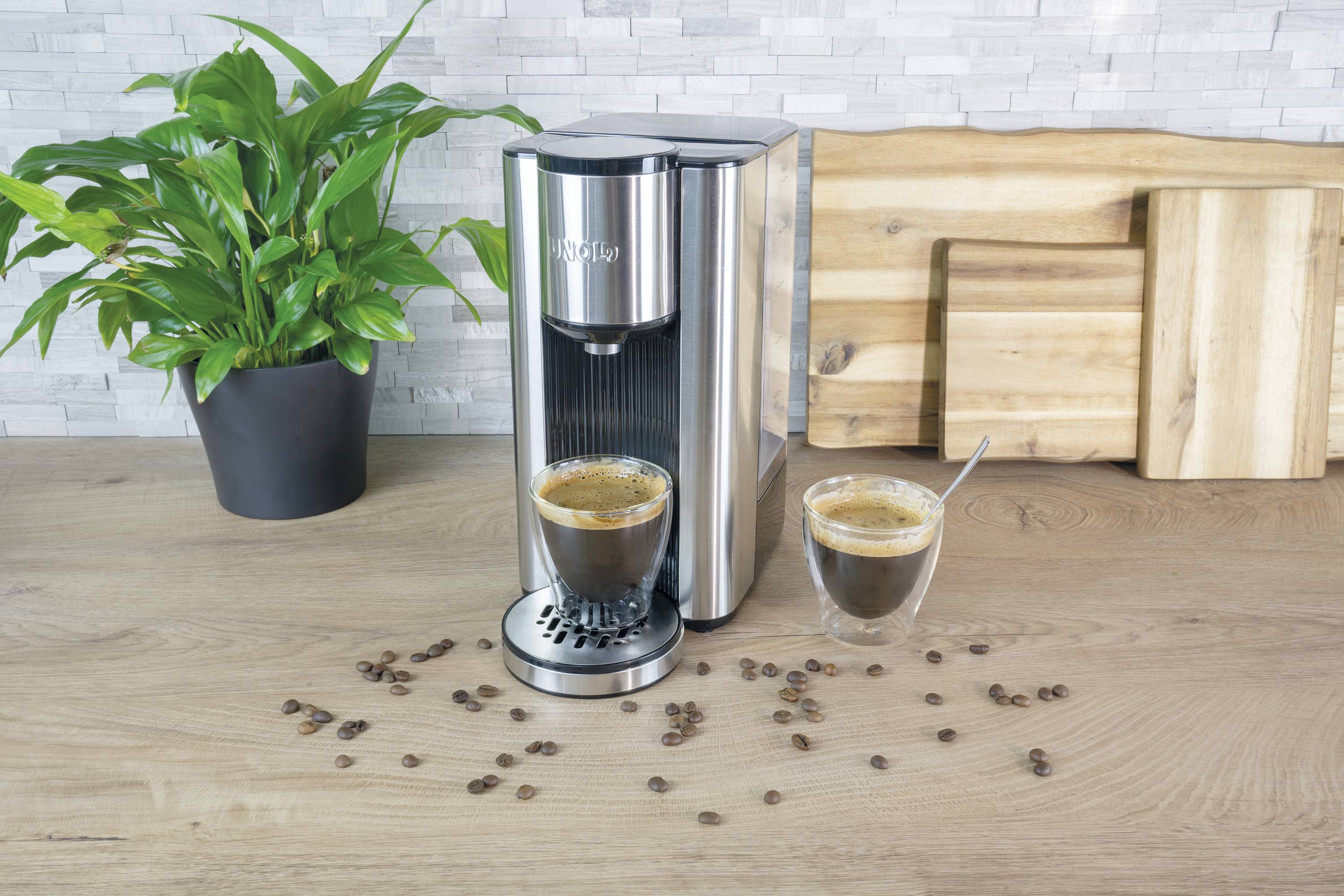 Elegancki ekspres do kawy stojący na blacie kuchennym z dwoma filiżankami pełnymi espresso; obok roślina doniczkowa i drewniane deski do krojenia.