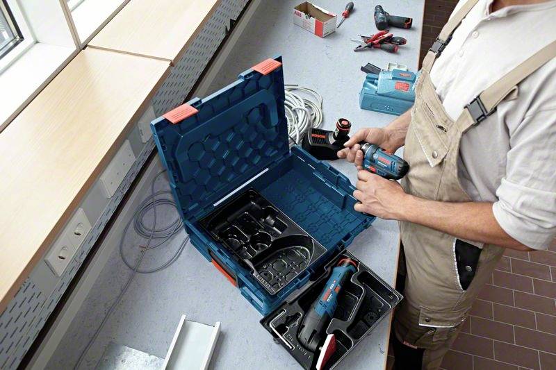 Osoba w stroju roboczym stoi przy stole z otwartą walizką narzędziową pełną elektronarzędzi i akcesoriów.