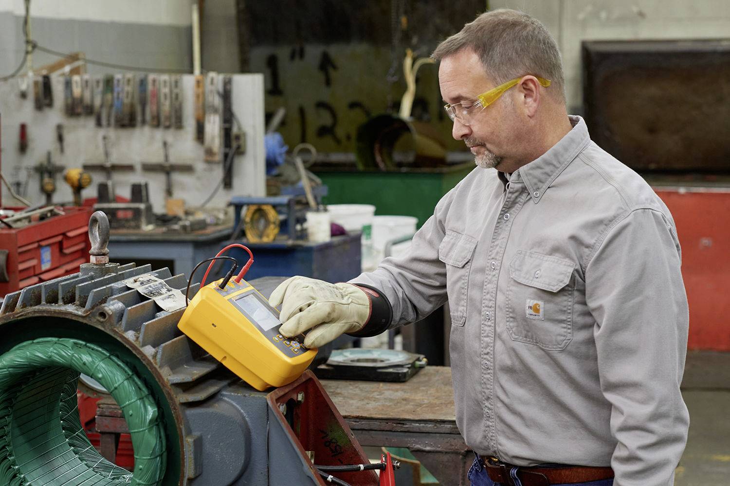 Mężczyzna w okularach ochronnych i rękawicach sprawdza dużą maszynę za pomocą urządzenia pomiarowego w warsztacie.