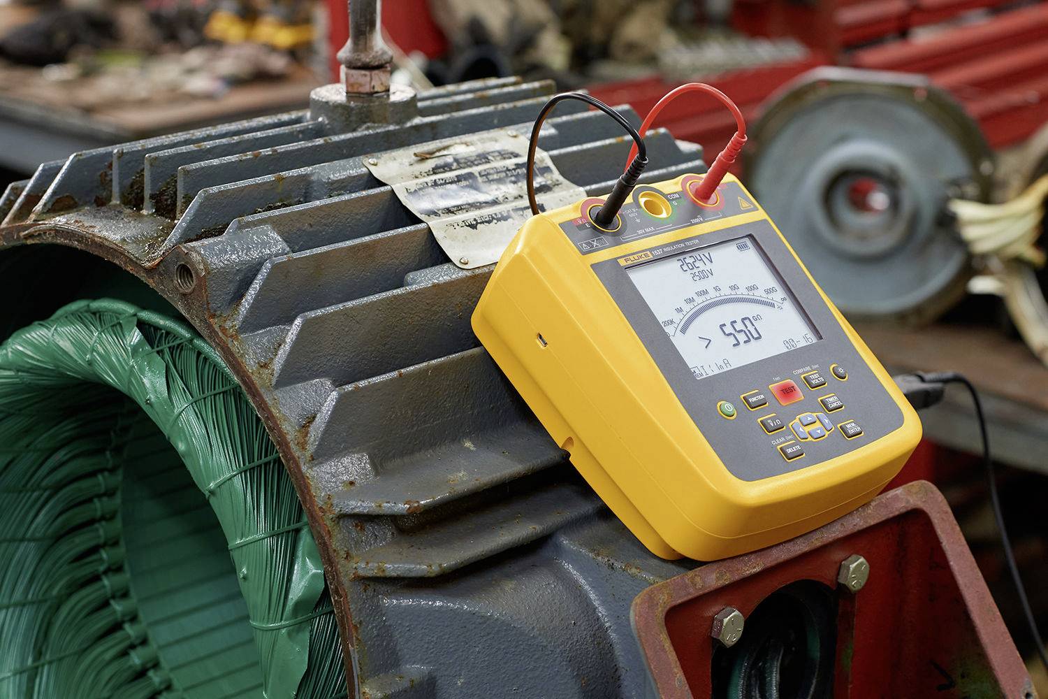 Żółte elektryczne urządzenie pomiarowe z wyświetlaczem pokazuje '550V' i leży na dużym, otwartym silniku elektrycznym. Otoczenie to warsztat.