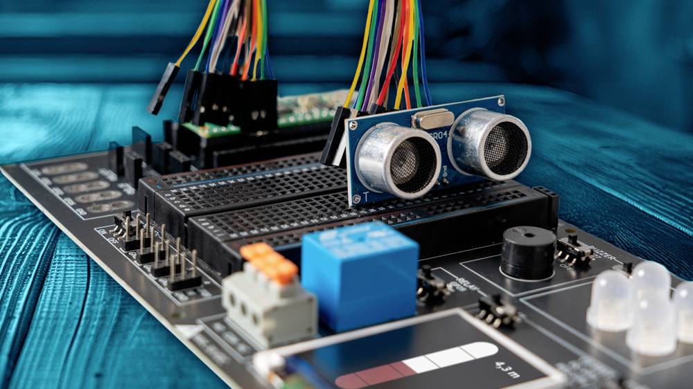 Płytka elektroniczna z czujnikami i kolorowymi przewodami na stole, prezentująca komponenty technologiczne do projektów majsterkowiczów lub programowania.