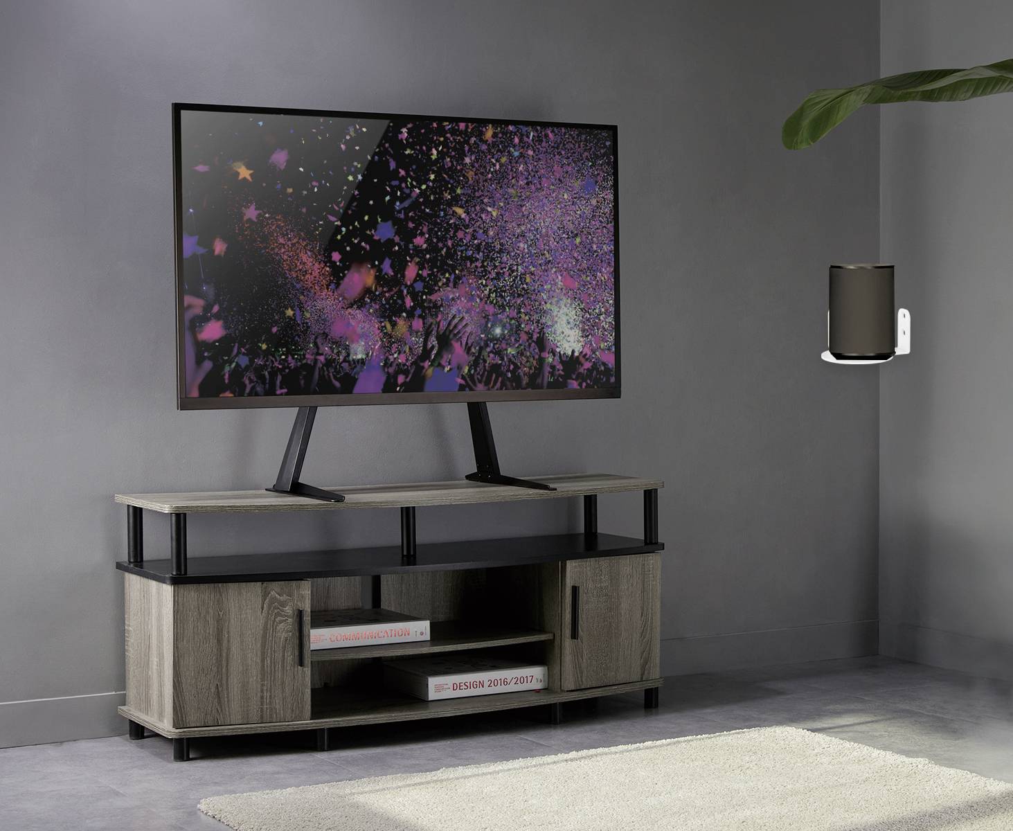 Telewizor stojący na drewnianej szafce wyświetla kolorowe sceny konfetti. Obok niego na ścianie zamontowany jest mały głośnik.