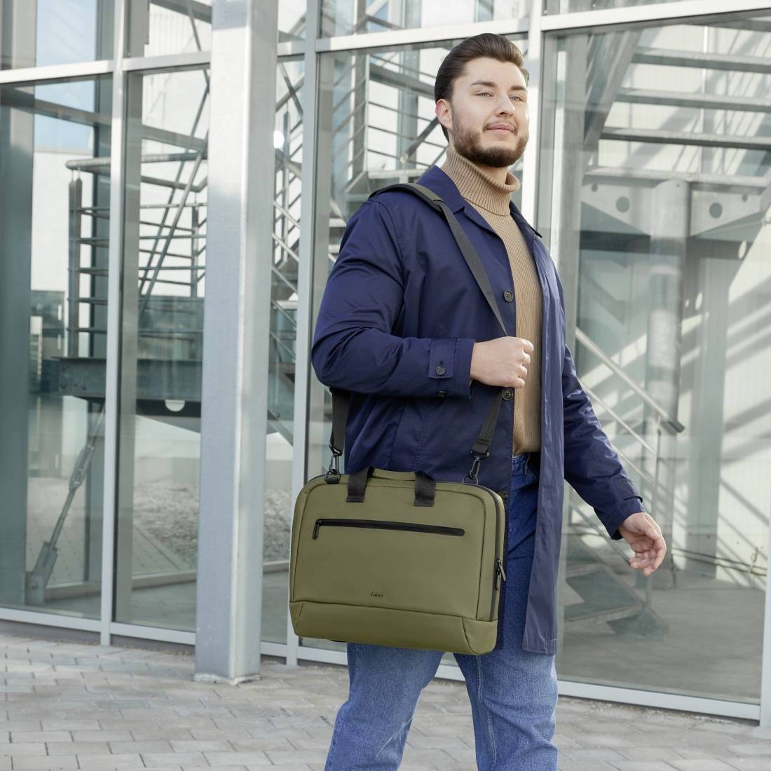 Mężczyzna stoi przed nowoczesnym budynkiem ze szklaną fasadą. Ma na sobie płaszcz, trzyma oliwkowozieloną torbę i patrzy pewnie.