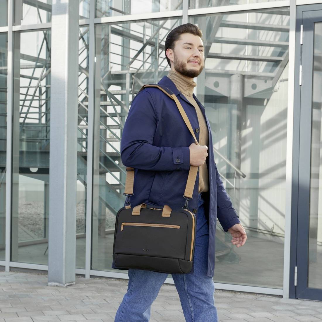 Mężczyzna stoi na zewnątrz przed nowoczesnym budynkiem, ubrany w ciemnoniebieską kurtkę, brązowy golf i trzyma czarną teczkę.