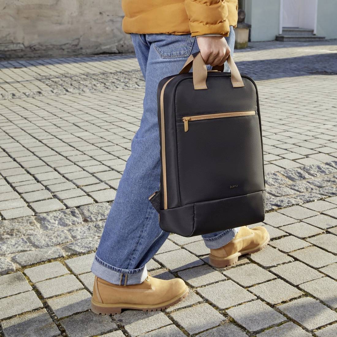 Osoba ubrana w niebieskie dżinsy i żółtą kurtkę nosi czarny plecak i brązowe buty, idąc po wybrukowanej ścieżce.