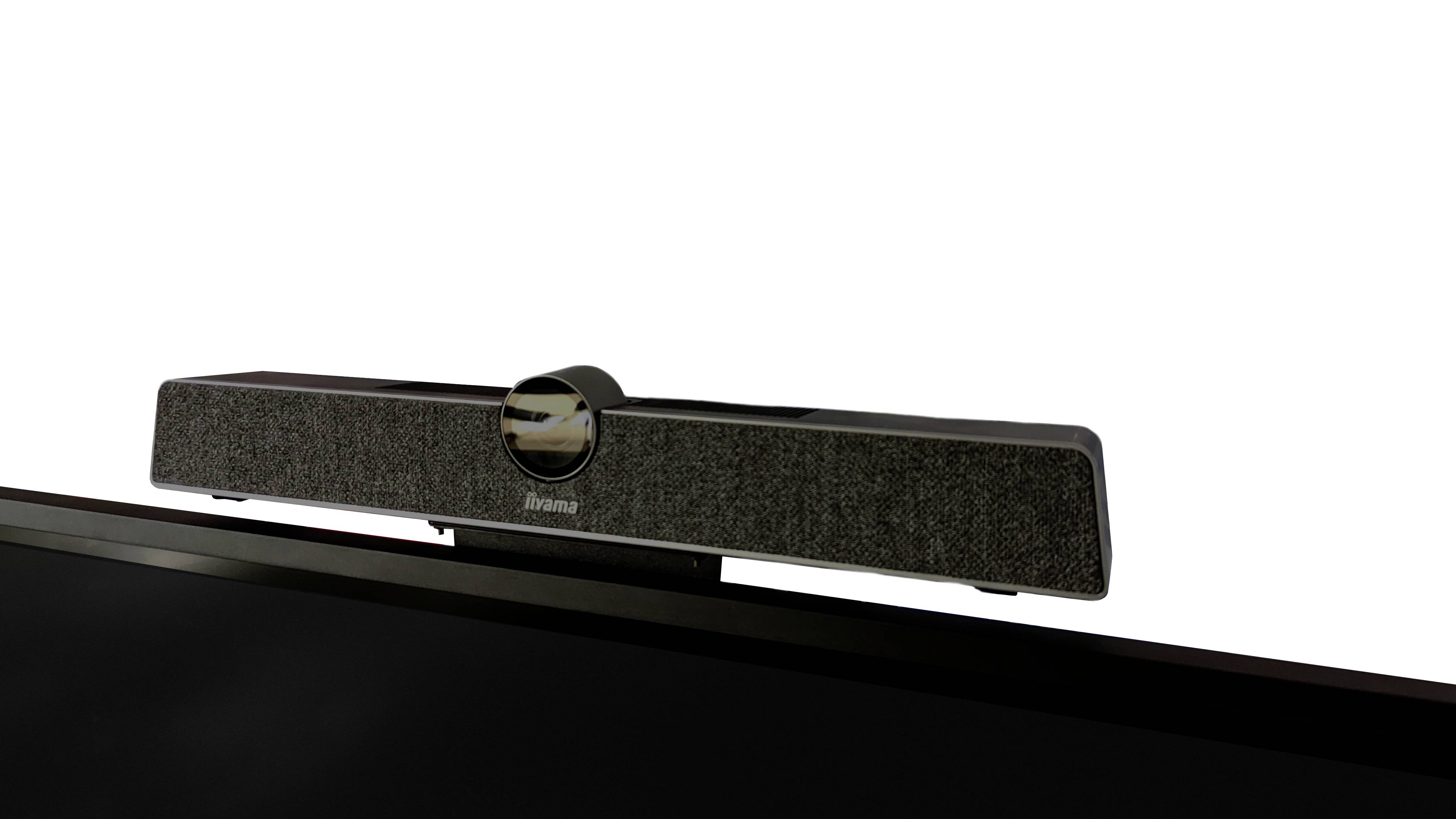 Soundbar spoczywa na monitorze, jej przednia strona jest pokryta tkaniną, a pośrodku widać błyszczące, okrągłe logo.