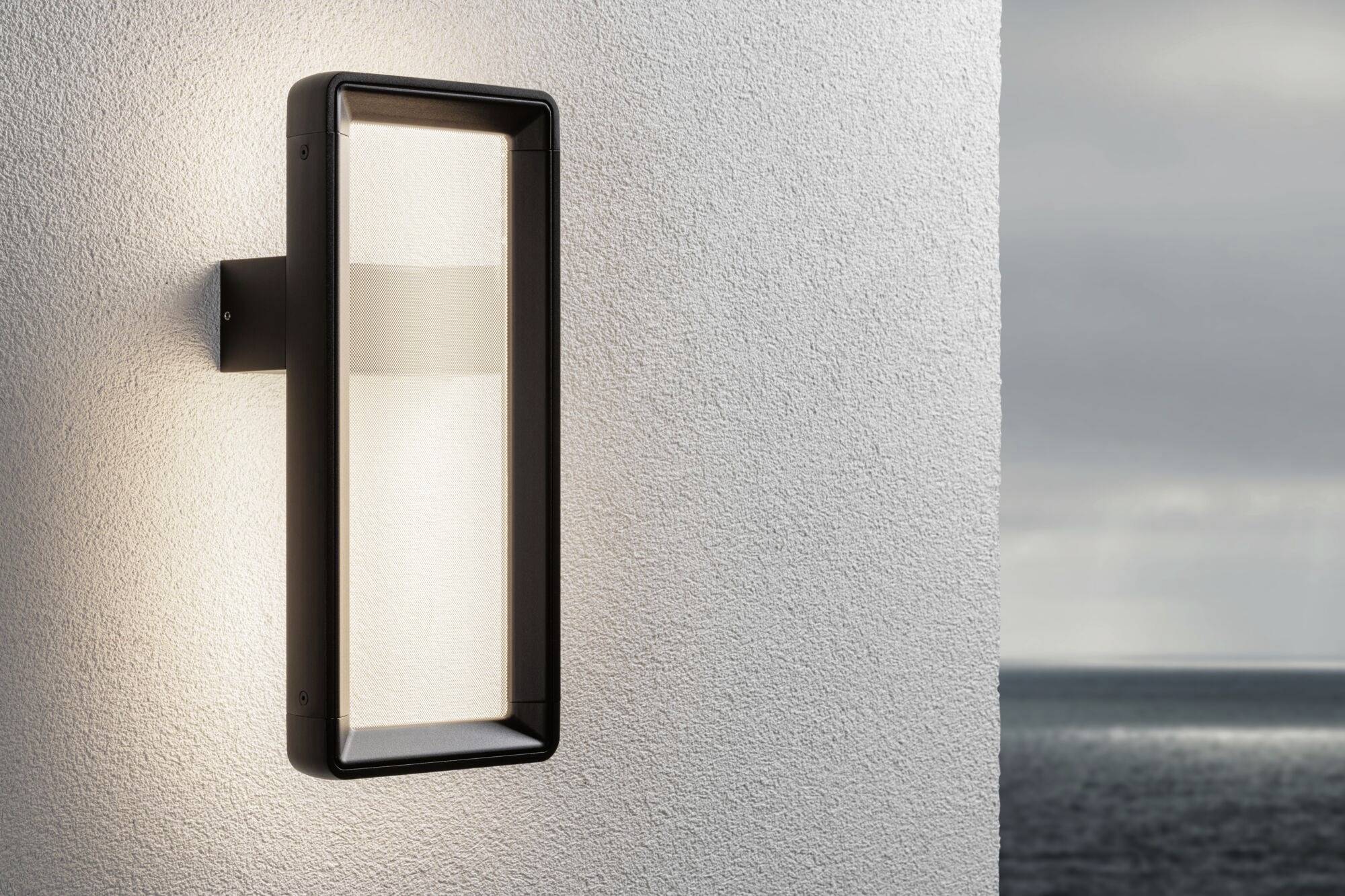 Lampa zewnętrzna świeci na białej ścianie, utrzymana w nowoczesnym, prostokątnym stylu. W tle ponury niebo rozciąga się nad morzem.