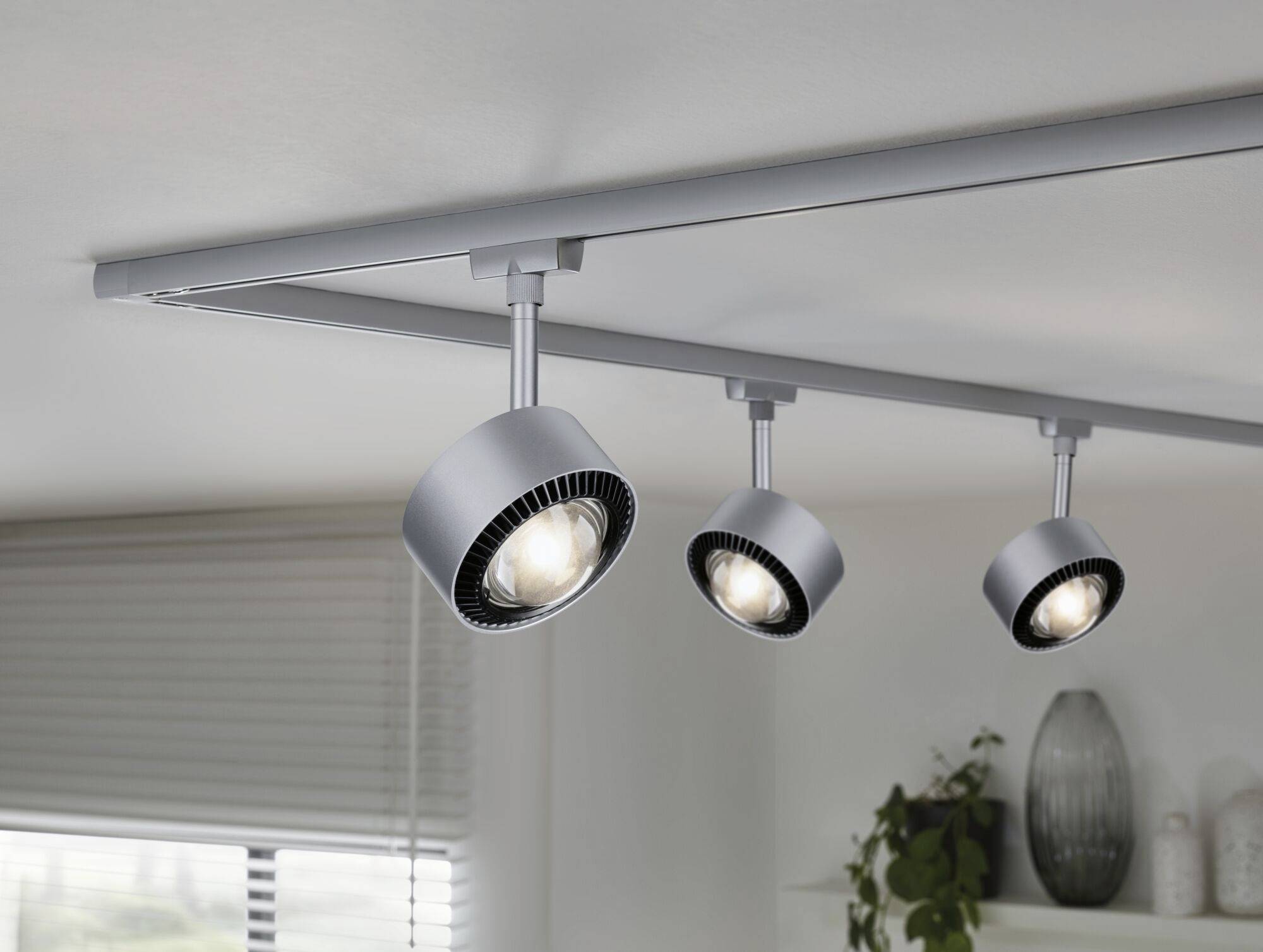 Lampa sufitowa z trzema regulowanymi reflektorami w nowoczesnym stylu, zamontowana na szynie w jasnym pomieszczeniu z roślinami w tle.