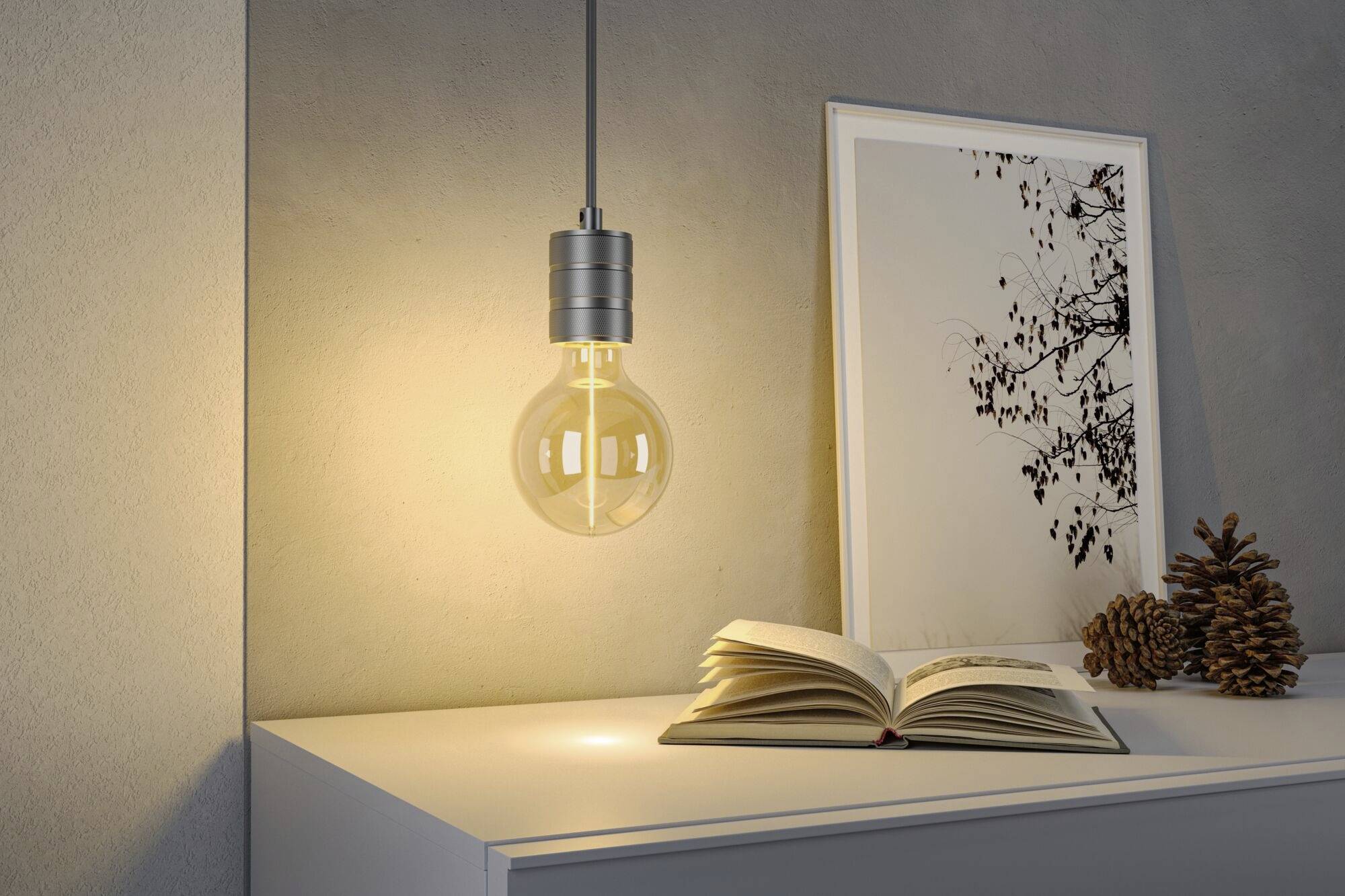 Świecąca żarówka wisi nad białym stołem z otwartą książką, obok znajduje się oprawiony obraz i szyszki.