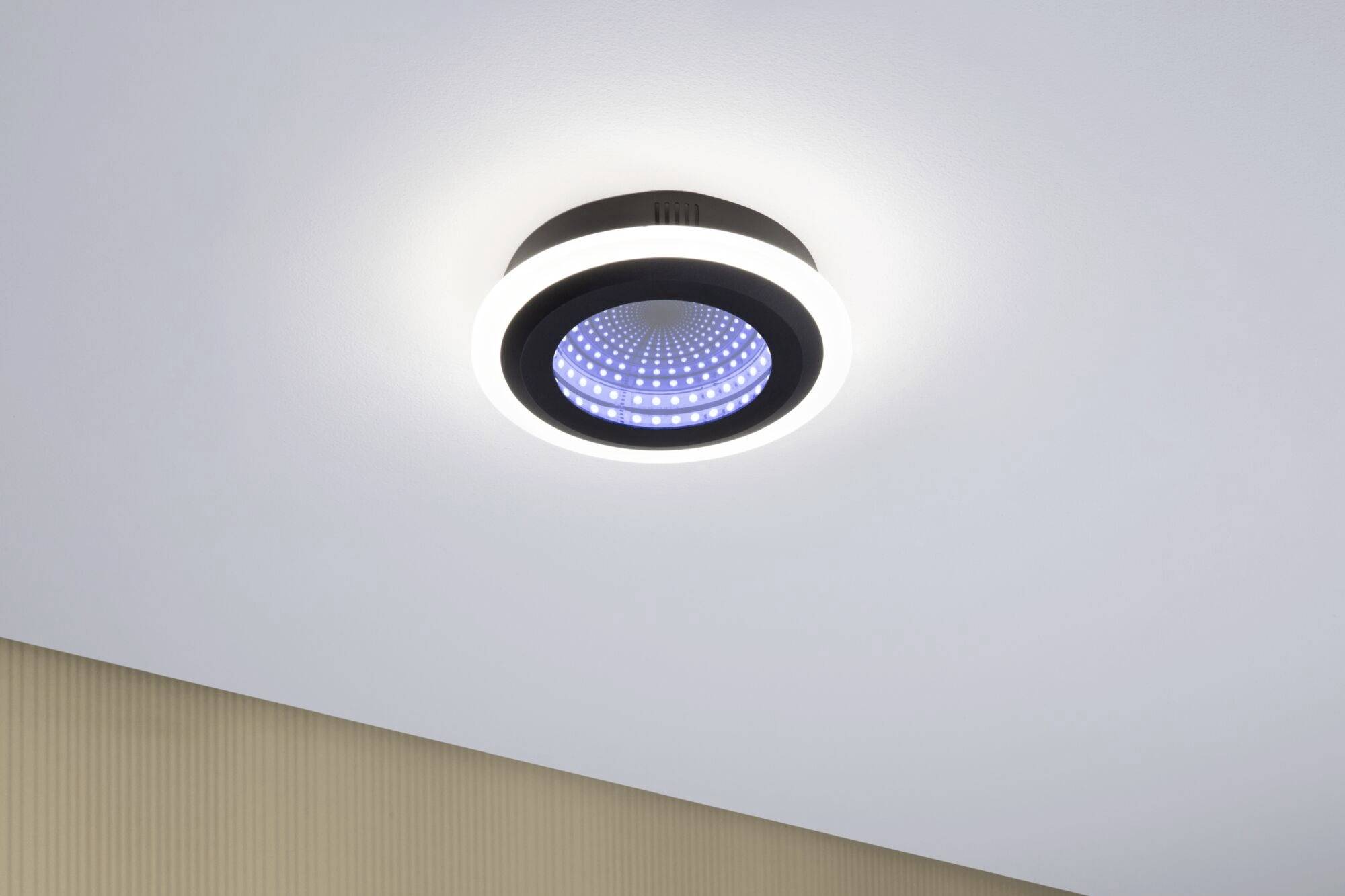 Nowoczesna lampa sufitowa emituje niebieskie i białe światło, zamontowana jest na białym suficie w pomieszczeniu o beżowych ścianach.