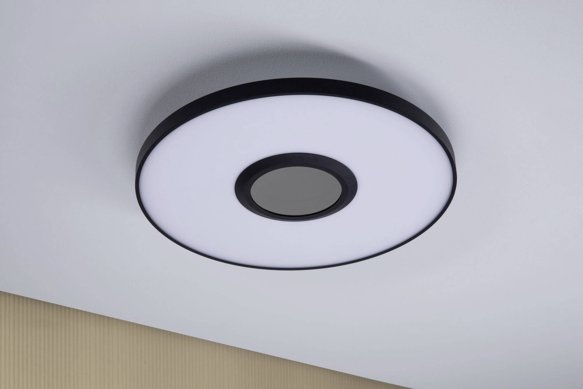 Okrągła nowoczesna lampa sufitowa z czarną obwódką, centralnym dyfuzorem i jasną powierzchnią świetlną, zamontowana na białym suficie.