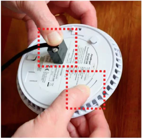 Osoba trzyma w ręku okrągły czujnik dymu, otwiera komorę baterii. Widoczny jest przewód. Urządzenie jest konserwowane.