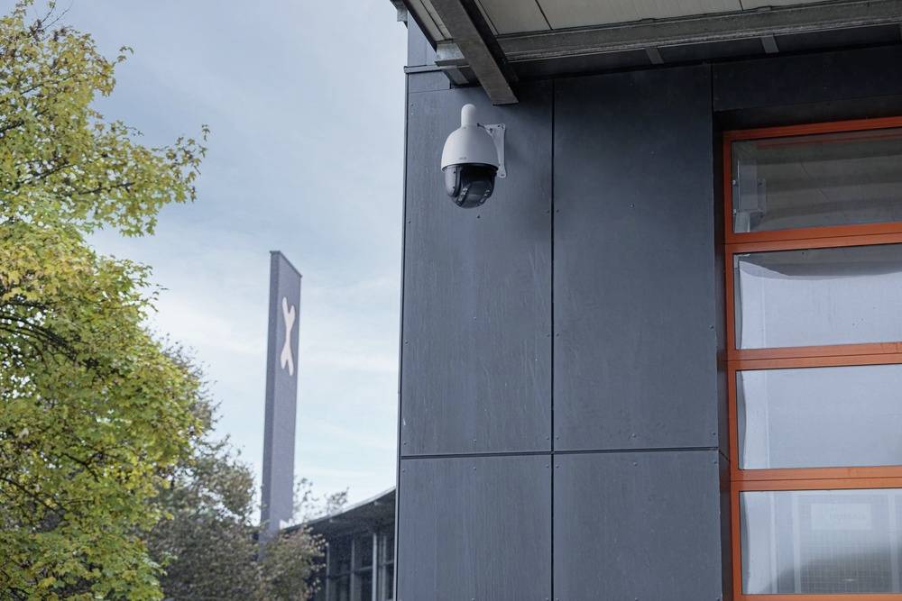 Kamera monitorująca jest zamontowana na zewnętrznej ścianie budynku. W tle widoczne są drzewa i znak.