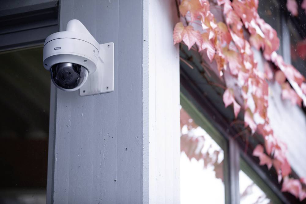 Kamera monitorująca na rogu budynku, obok pnącza o czerwonawych liściach. Ponury, zachmurzony nieb odbija się w oknie.