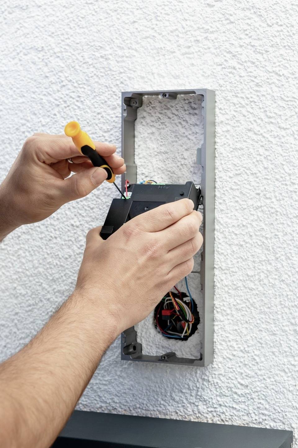 'Ktoś instaluje elektryczne gniazdko w ścianie. Jedna ręka trzyma śrubokręt, druga przytrzymuje przewody.'