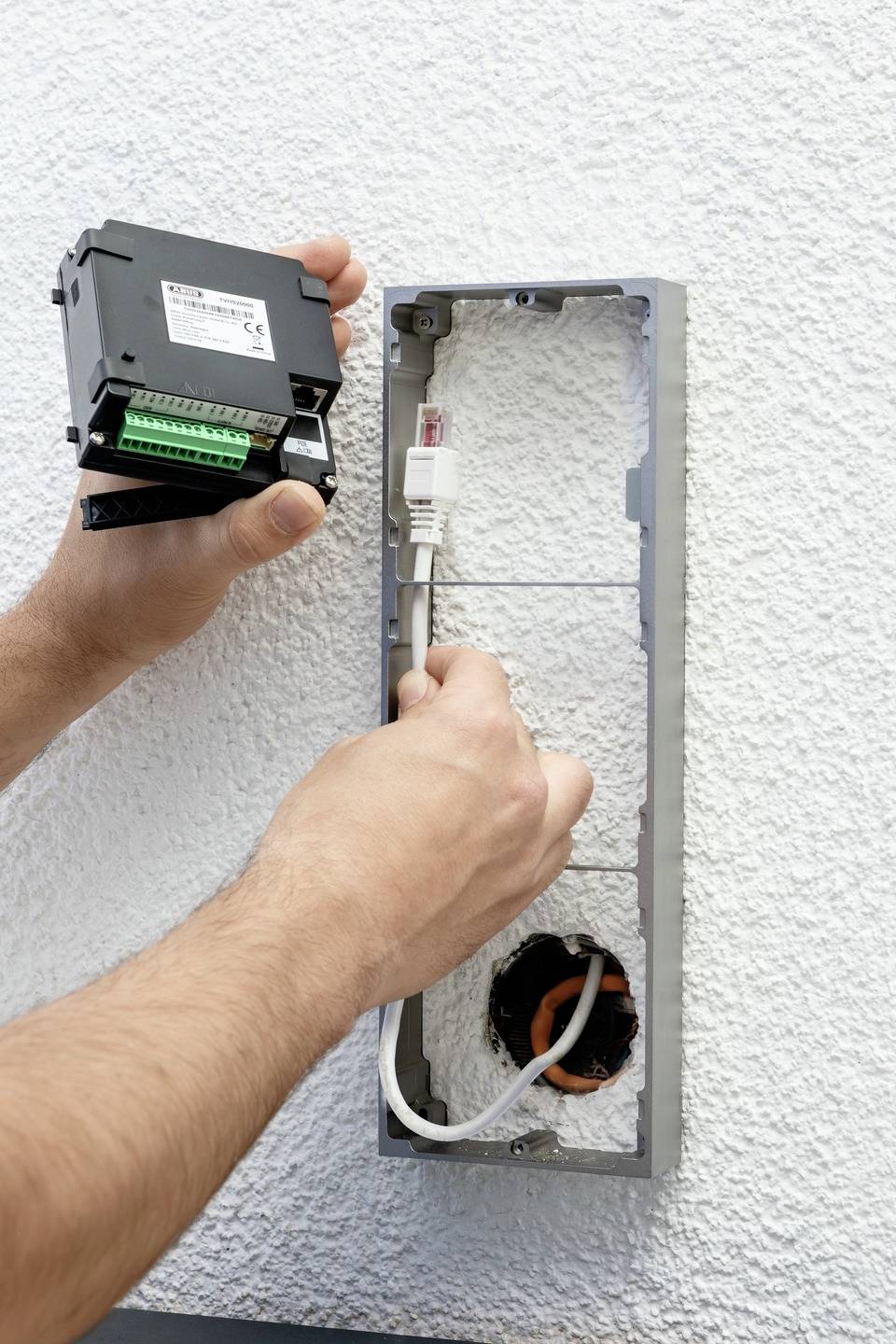 Osoba instaluje urządzenie elektroniczne w puszce ściennej. Widoczny jest kabel ethernetowy oraz dodatkowy przewód.