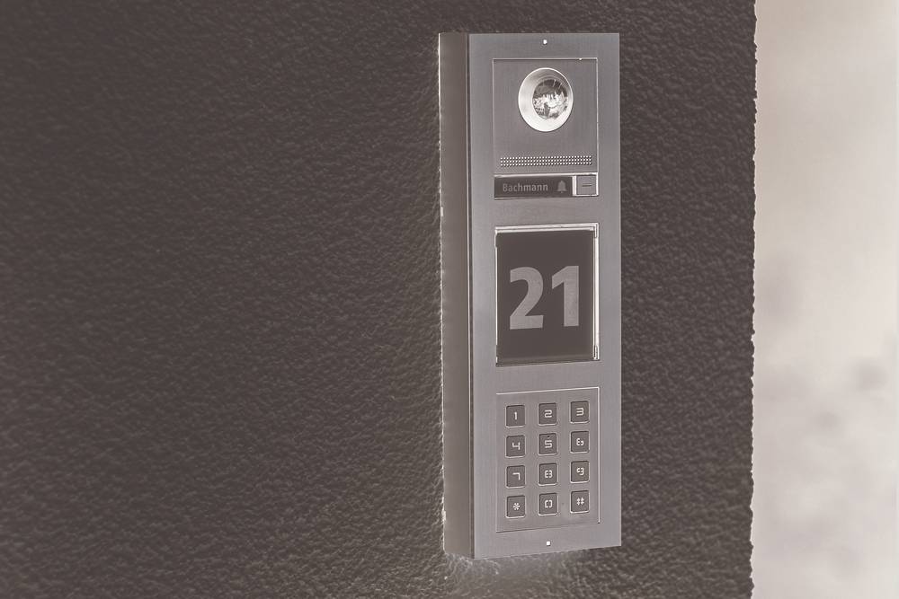Nowoczesny domofon z kamerą, głośnikiem i klawiaturą numeryczną jest zamontowany na ciemnej ścianie i wyświetla numer '21'.