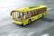 Żółty plastikowy autobus modelowy jedzie po drodze. Autobus ma zielone koła i przezroczyste okna.