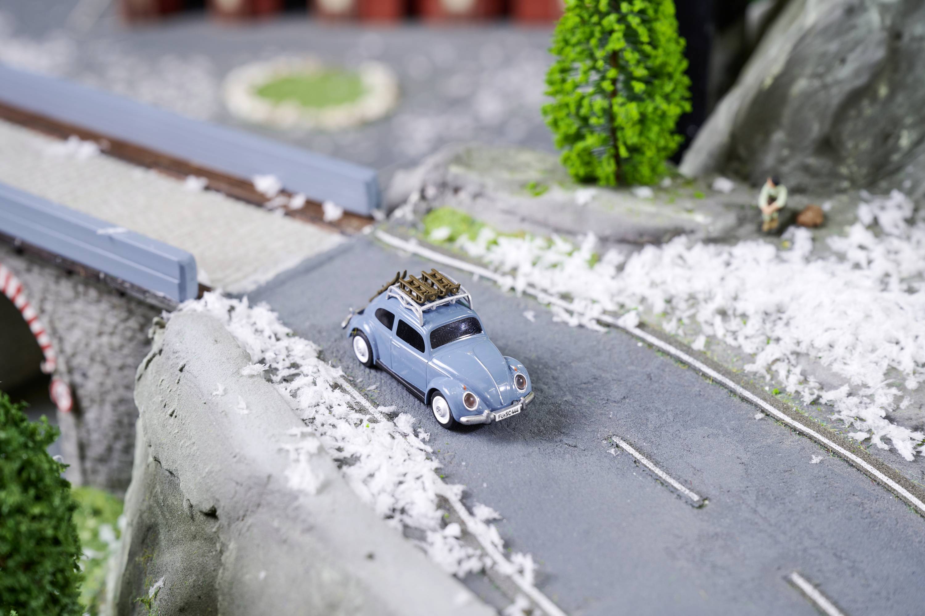 Miniaturowy samochodzik jedzie po małej drodze przez most. W tle widać modelowe drzewa i szczegóły krajobrazu.