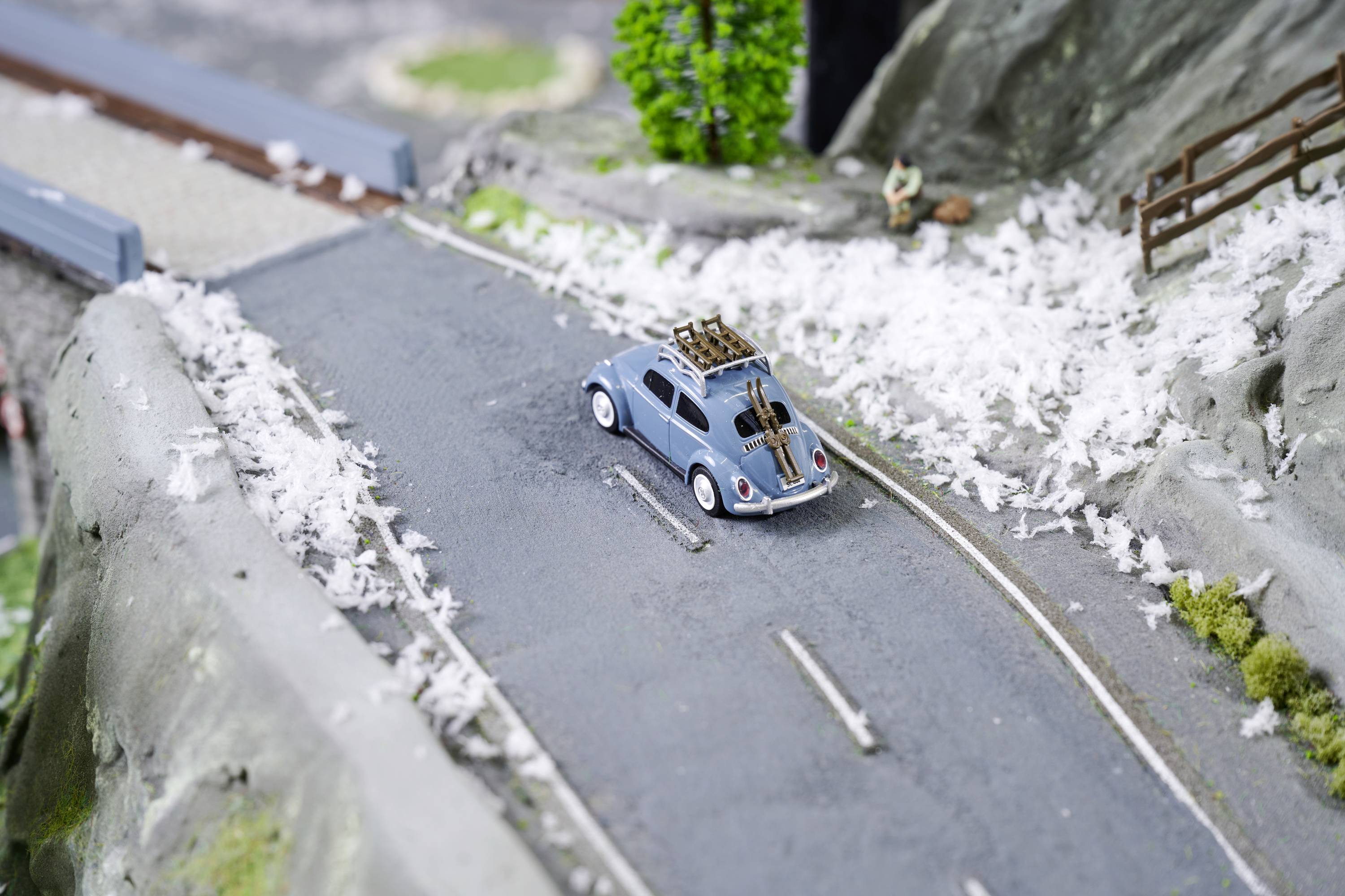 Miniaturowy samochód na zaśnieżonej górskiej drodze, z nartami przewożonymi na dachu. W tle widoczny płot i drzewa.