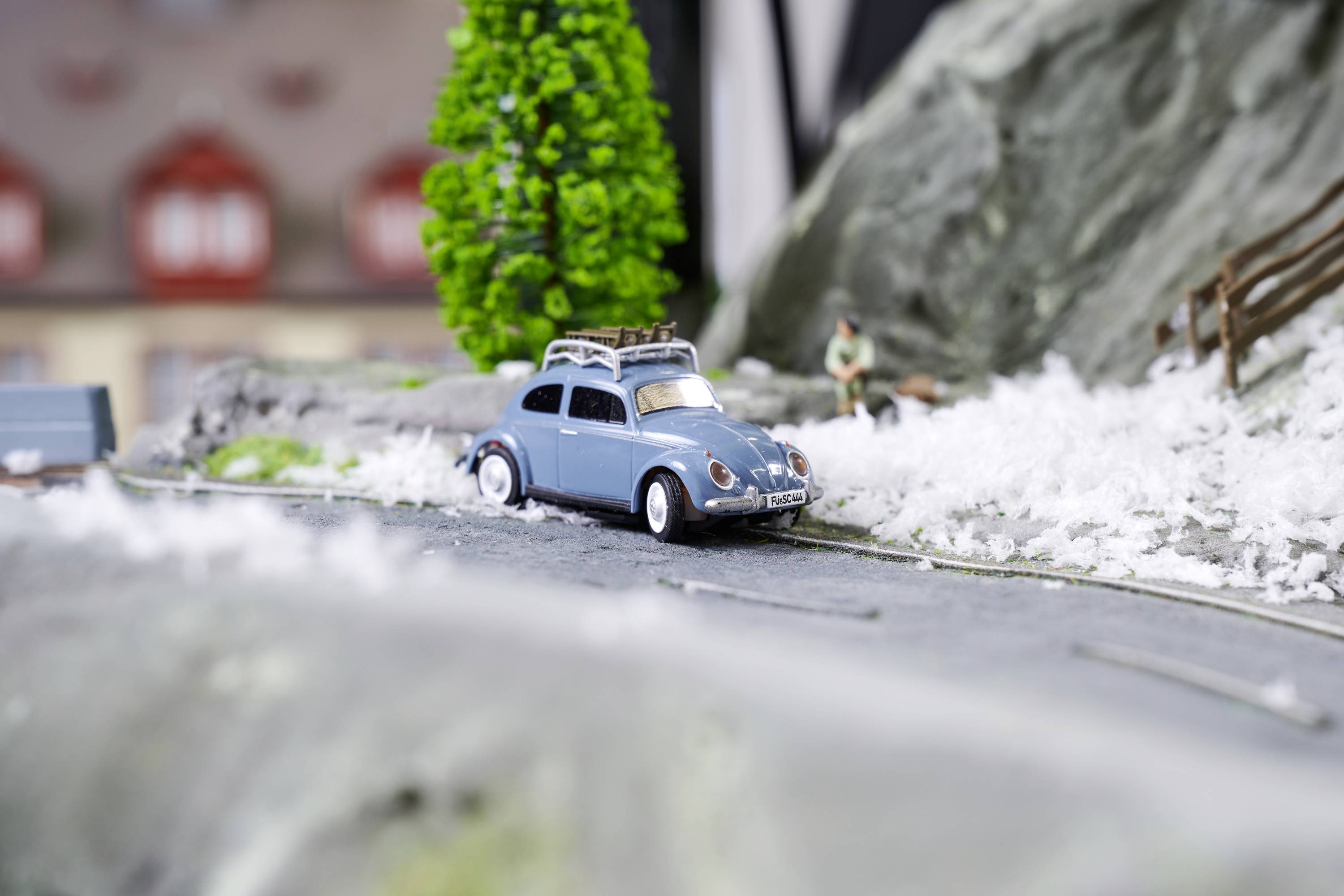 Niebieskie autko zabawkowe jedzie wzdłuż zaśnieżonej drogi. W tle stoi mały drzewiasty, a miniaturowa figurka biegnie.