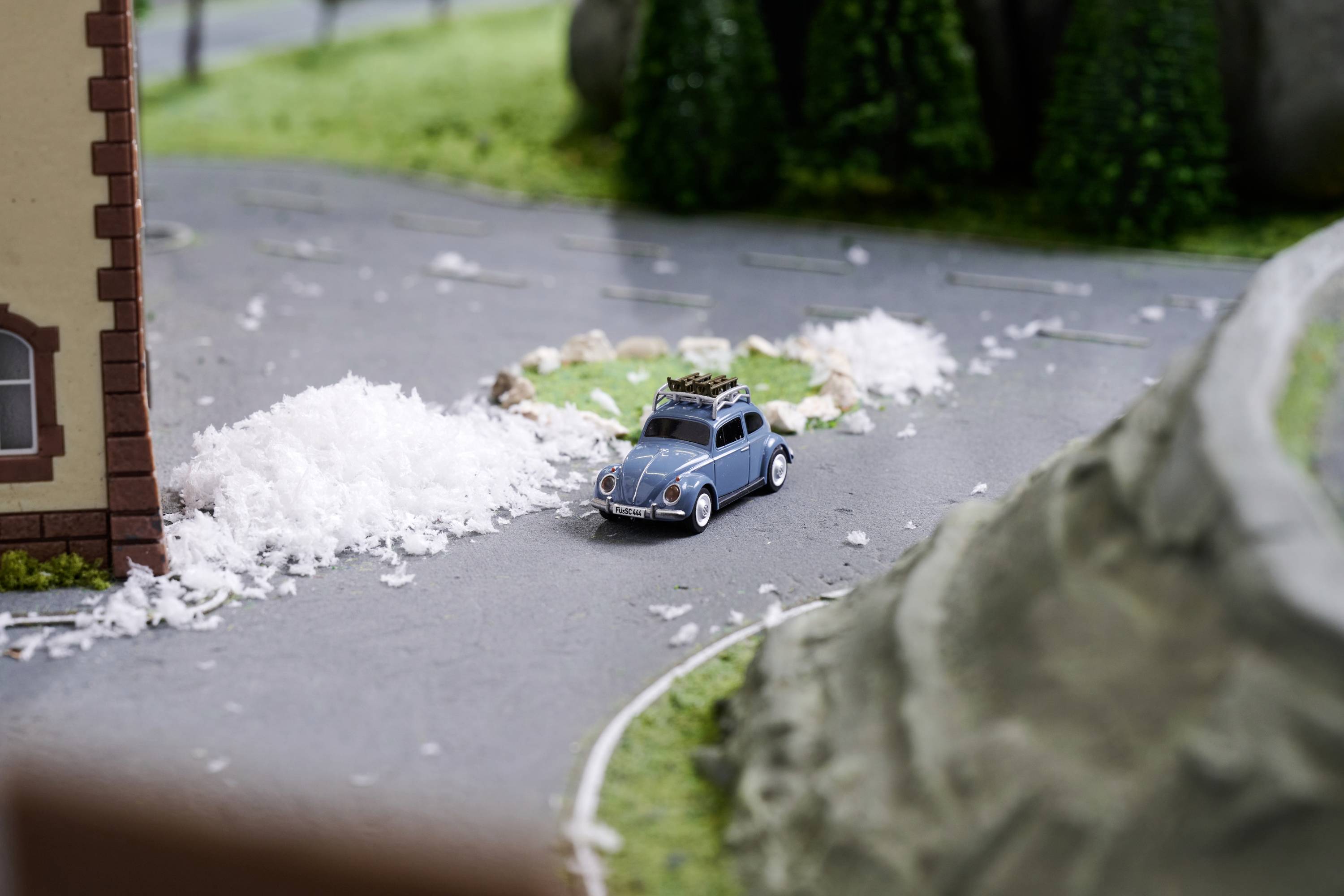 Zabawkowy samochodzik jedzie po modelowej drodze otoczony sztucznym krajobrazem. Po prawej stronie wznosi się wzgórze, po lewej stoi budynek.