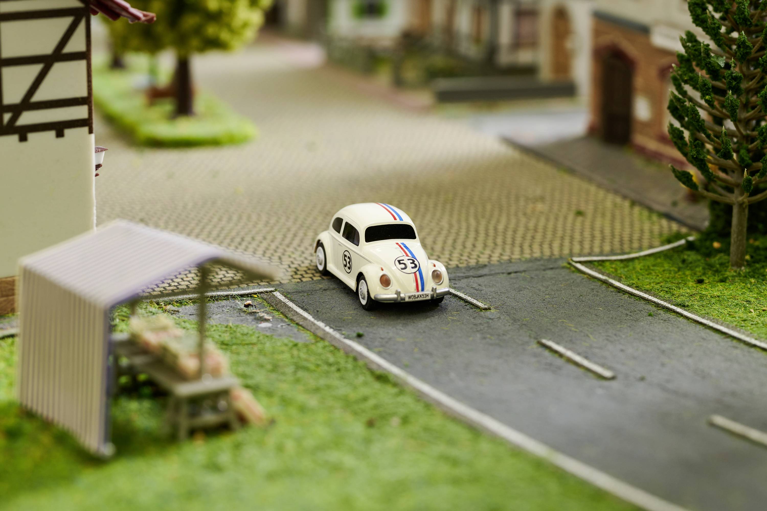 Mały biały samochodzik wyścigowy z pasami startowymi jedzie po krajobrazie makiety drogowej. W tle widoczne są drzewa i budynki.