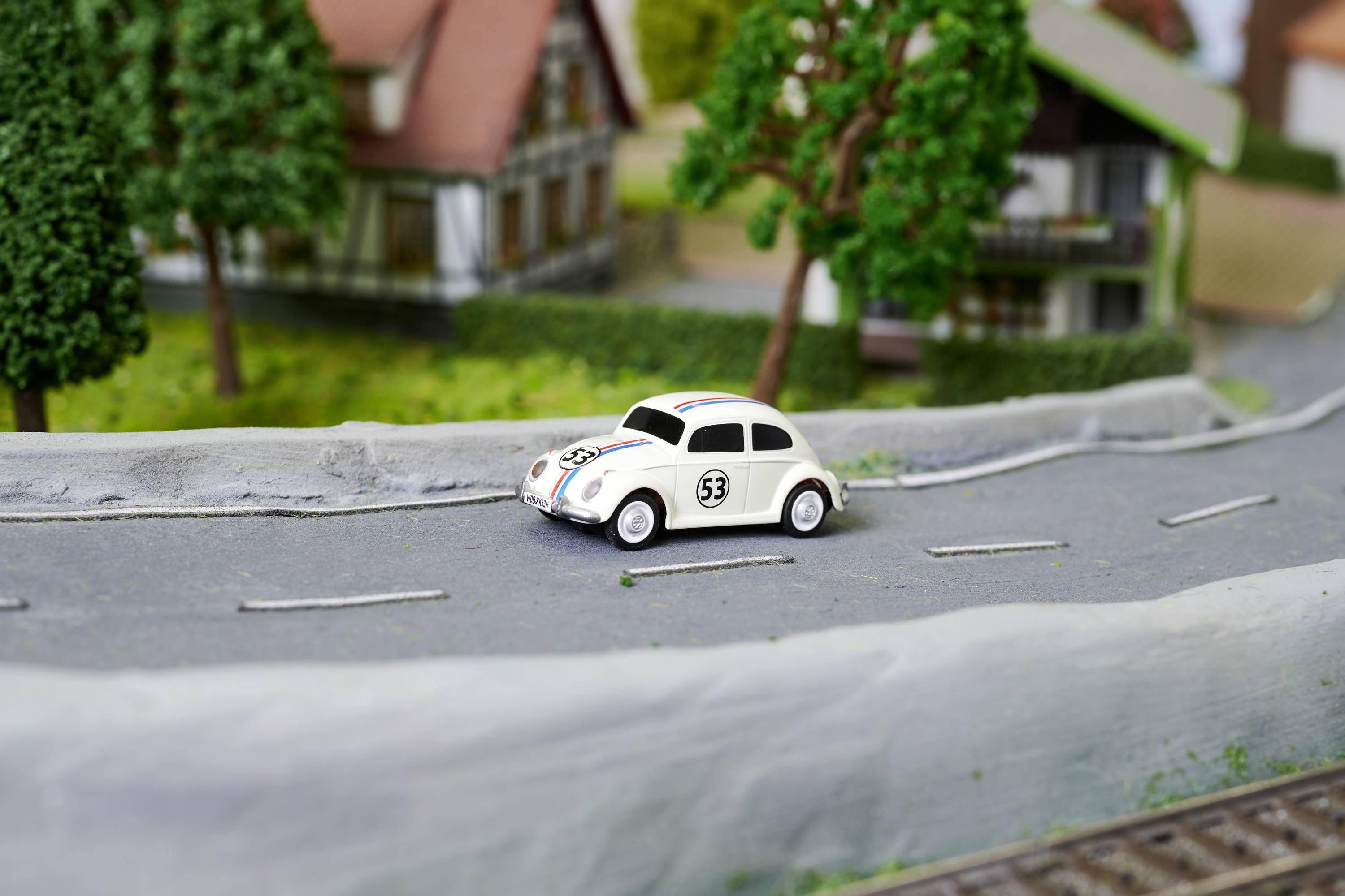 Mały biały samochodzik zabawkowy z numerem 53 jedzie po makiecie ulicy, mijając modelowane domy i drzewa.