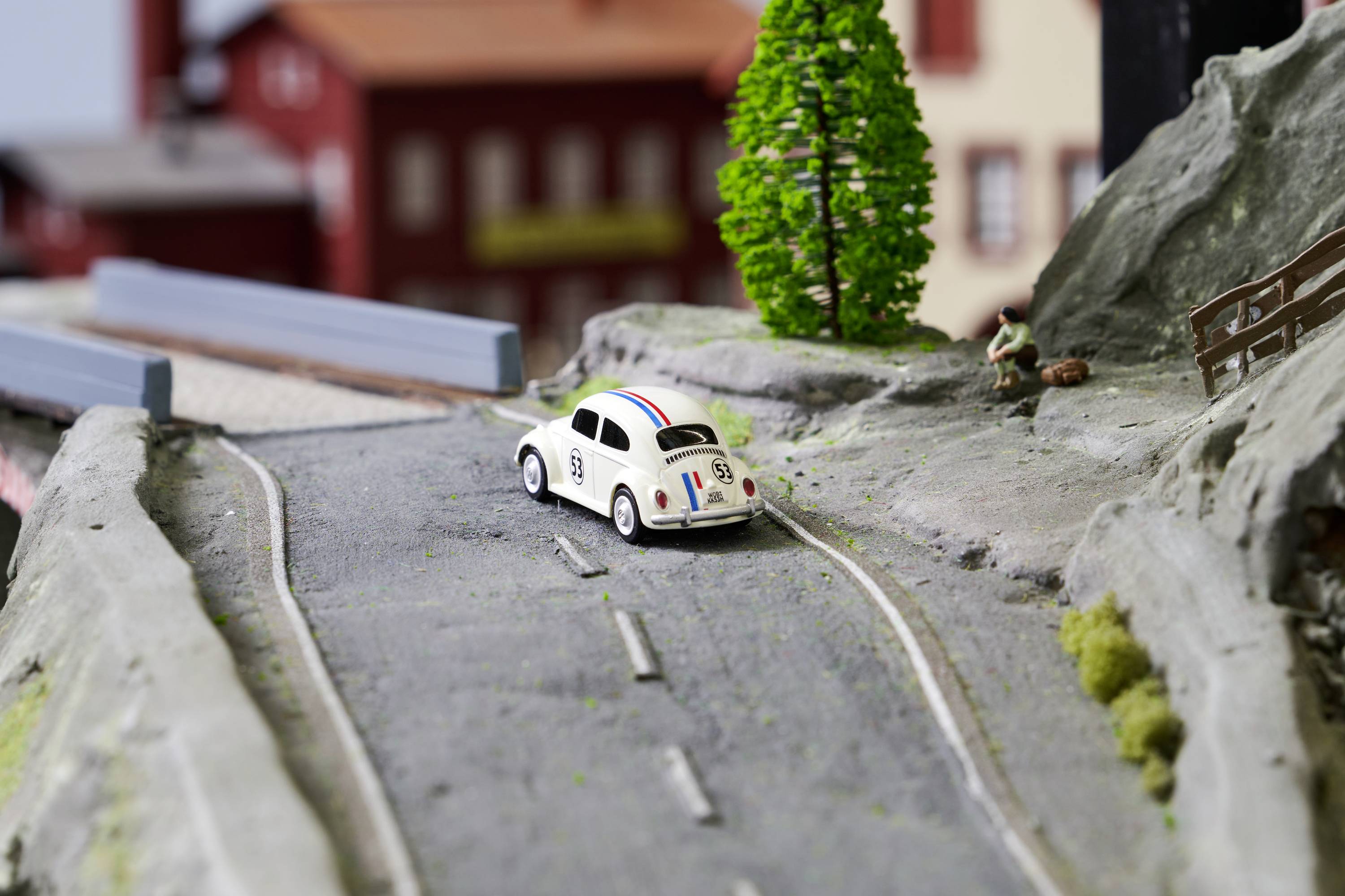 Mały samochodzik zabawkowy jedzie po miniaturowej drodze w modelu makiety; dziecko siedzi przy jej krawędzi.